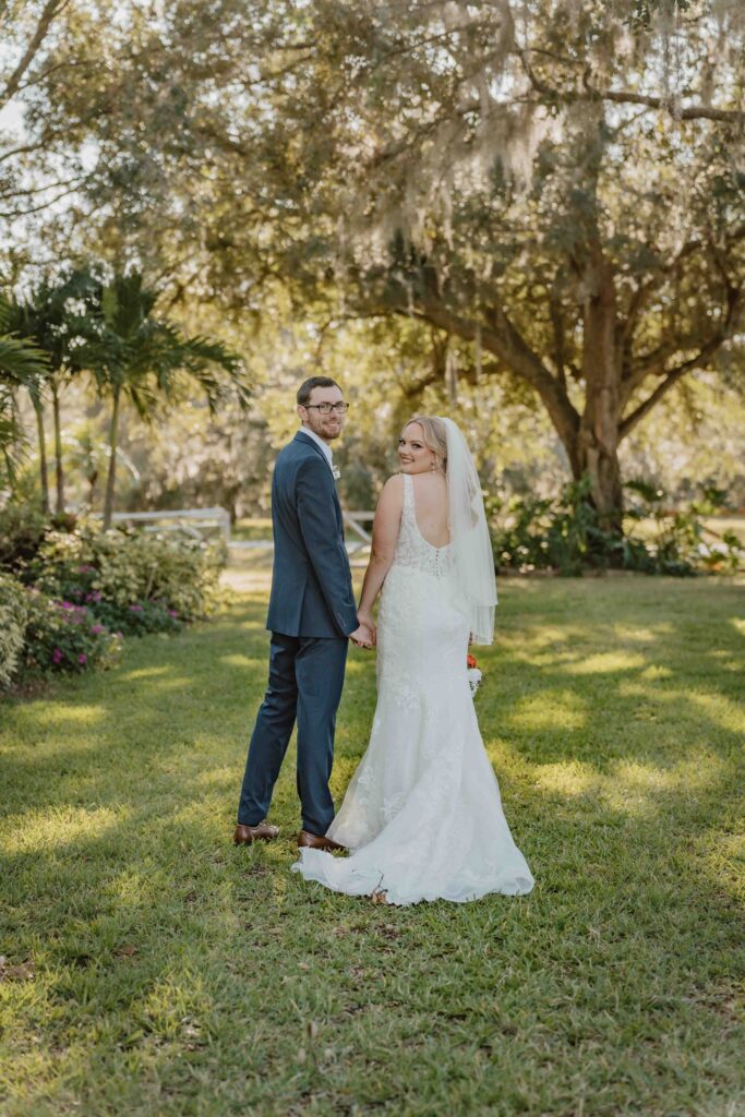 bride and groom holding hands in front of trees looking back at the camera