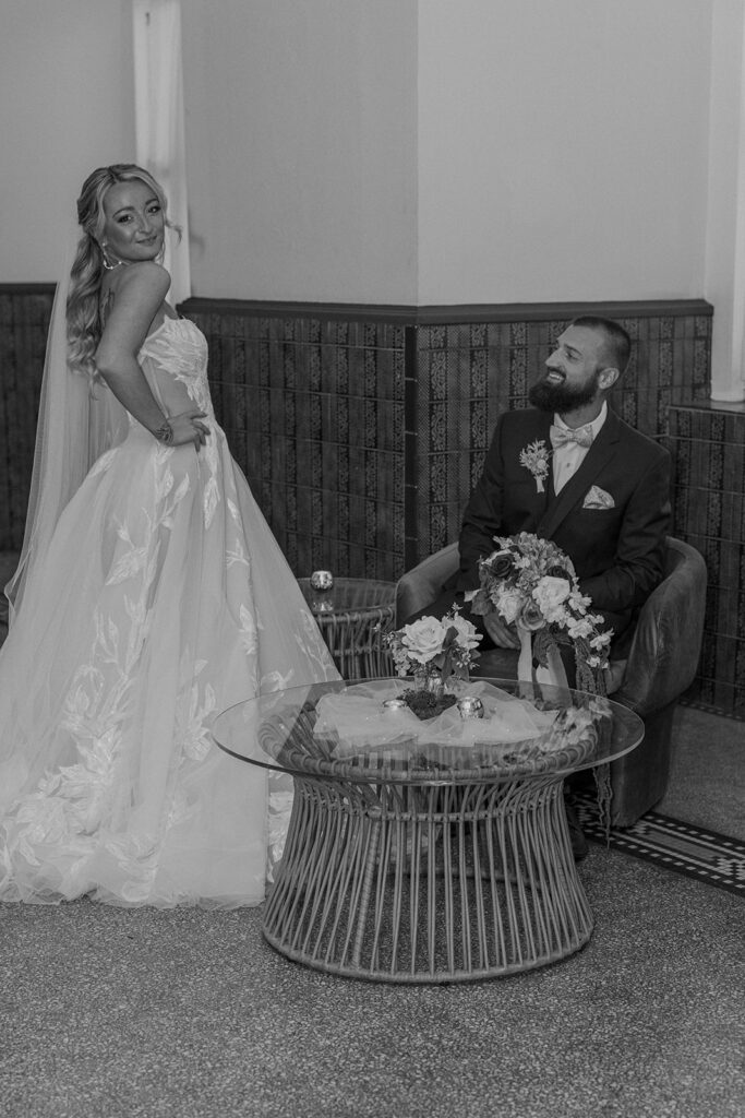 black and white photo of the bride standing and the groom sitting at cocktail hour