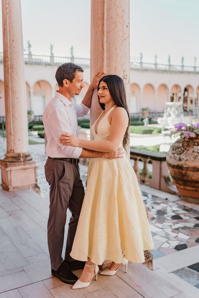 man and woman looking at each other with hands around her waits leaning against a column