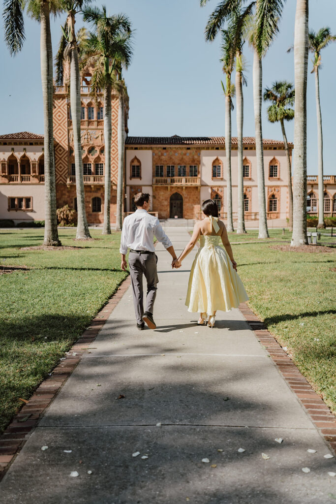 man and woman holding hands walking towards the museum on a sidewalk lined with palm trees