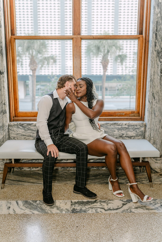 man and woman sitting on a bench in front of a window with him kissing her neck